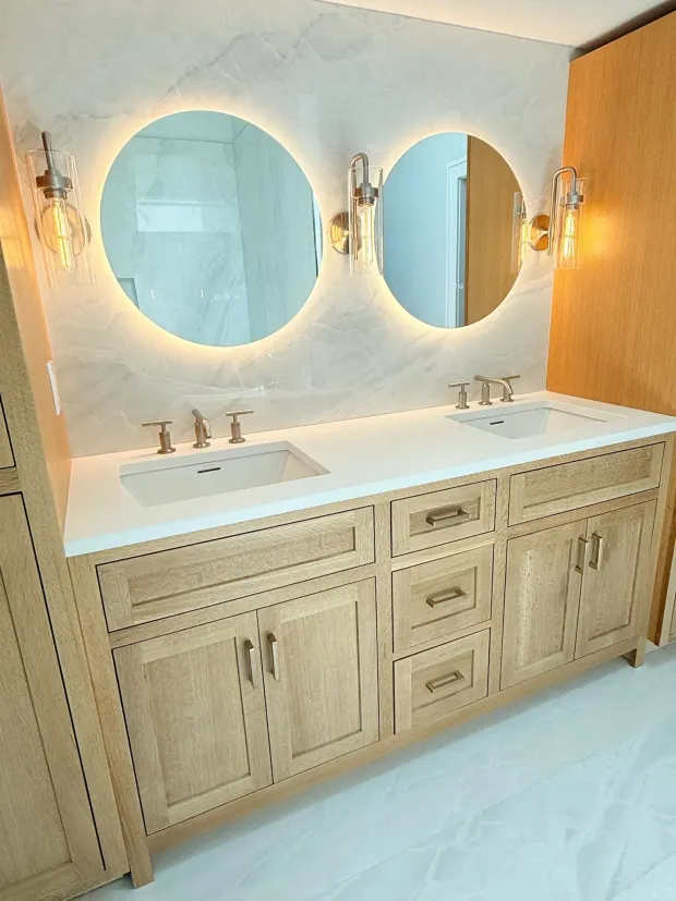 A bathroom with a light wood double vanity, two round illuminated mirrors, and sconces.