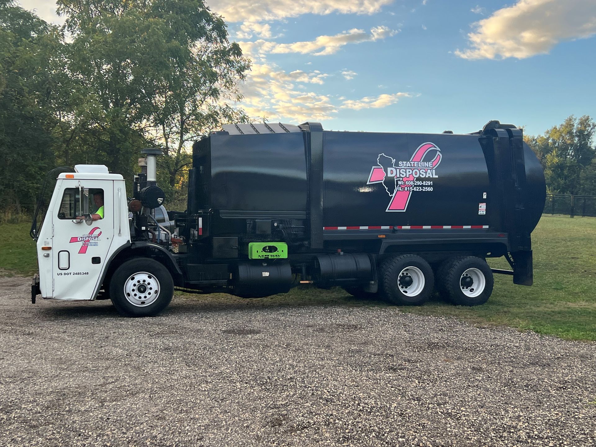 A black garbage truck with a pink ribbon on the side is parked in a gravel lot