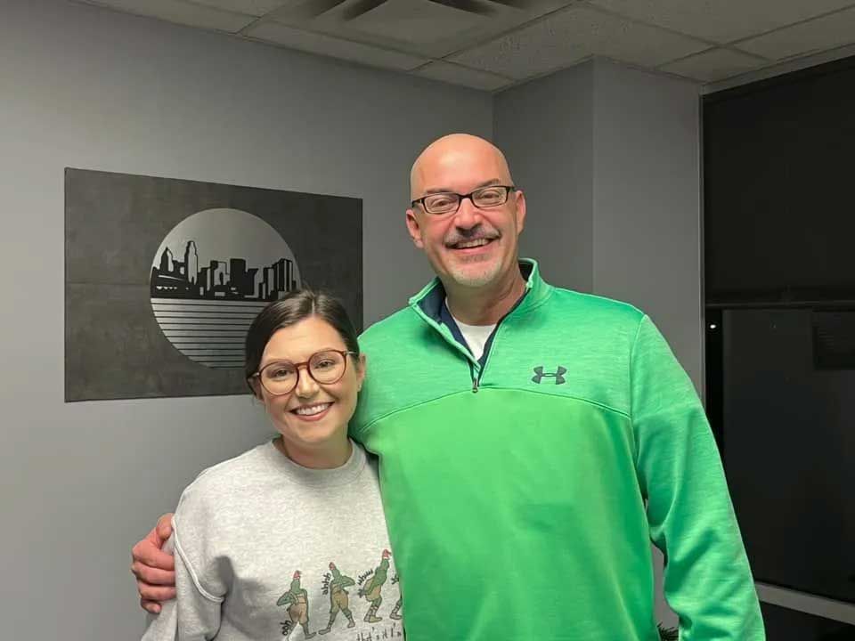 Two people smiling and posing, one with a hand on the other's shoulder, in an office. A cityscape art piece is on the wall.