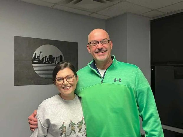 Two people smiling and posing, one with a hand on the other's shoulder, in an office. A cityscape art piece is on the wall.