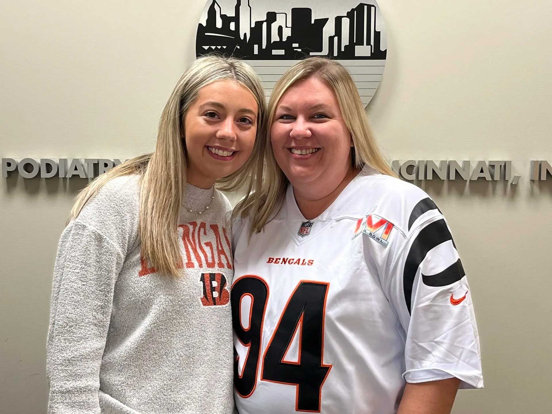 Two women smiling, posing in front of "Podiatry of Cincinnati, Inc." sign, one wearing a Bengals jersey.