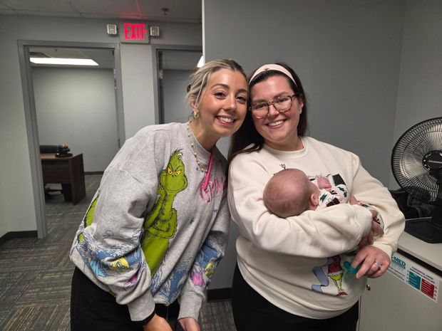 Two women smiling; one holds a baby. Office setting. The woman on the left wears a Grinch sweater, the other a cream sweater.