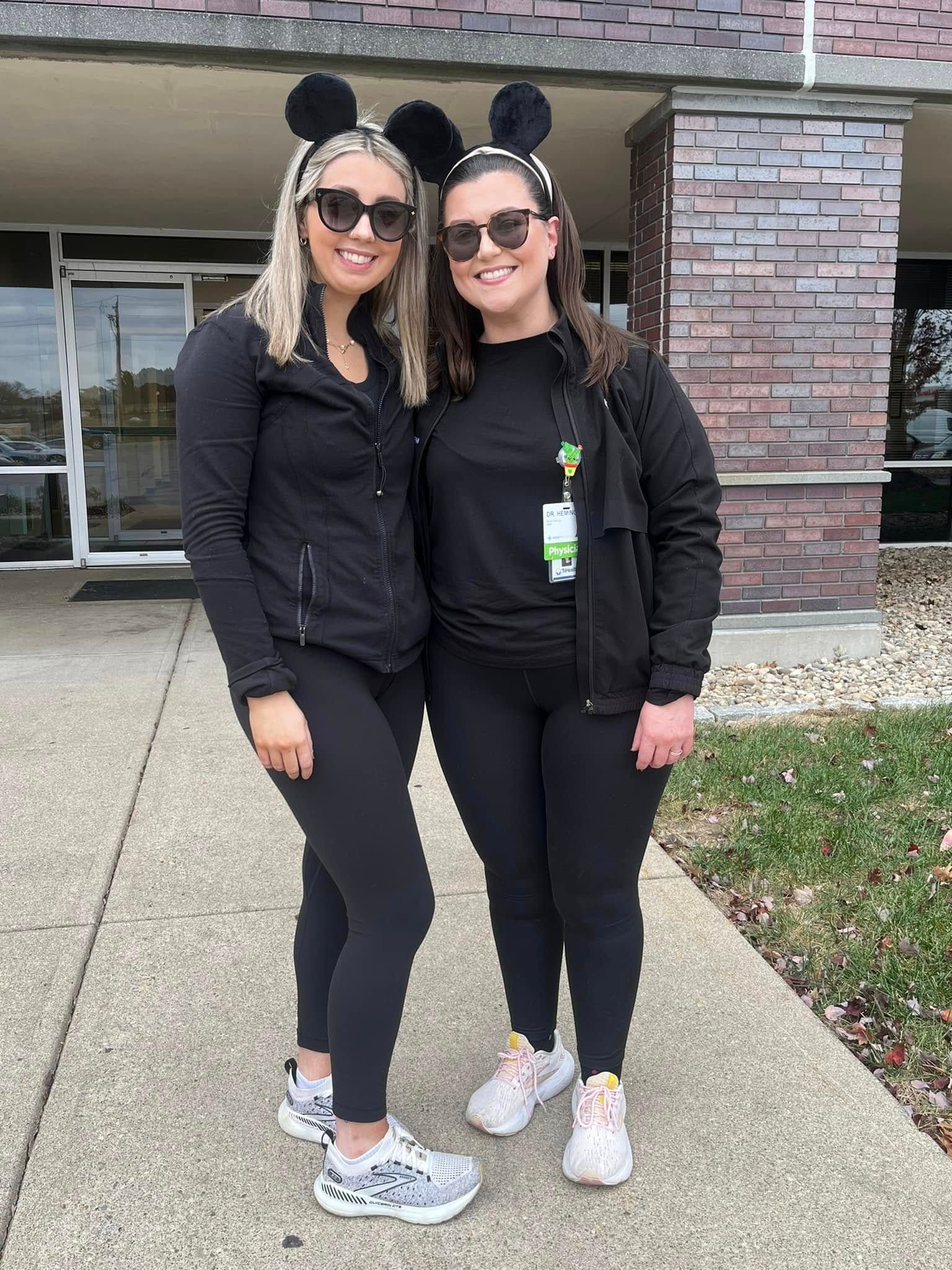 Two people wearing black outfits and ear headbands, outside building, smiling.