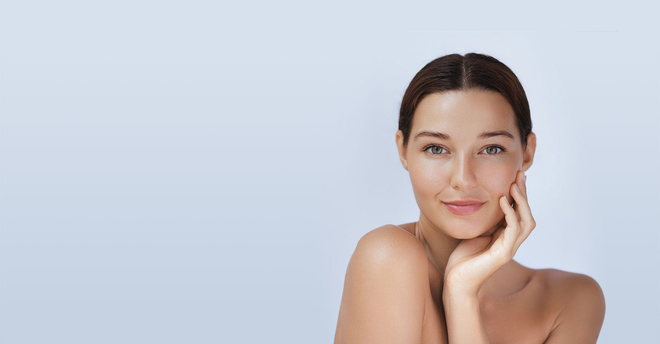 Woman with clear skin touching her face, smiling; light blue background.