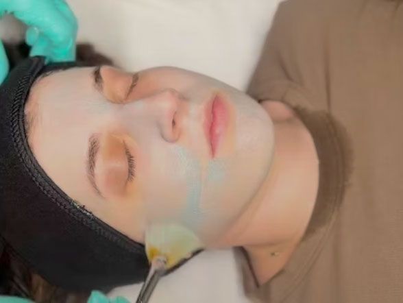 Woman with mask receiving facial treatment. Teal gloved hand uses a tool near cheek. Brown top, black headband.