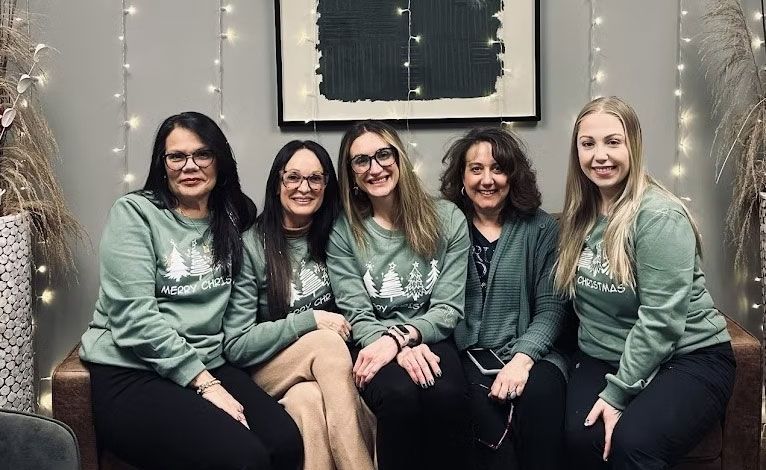 Five people wearing green sweaters, smiling, seated on a couch in front of artwork and string lights.