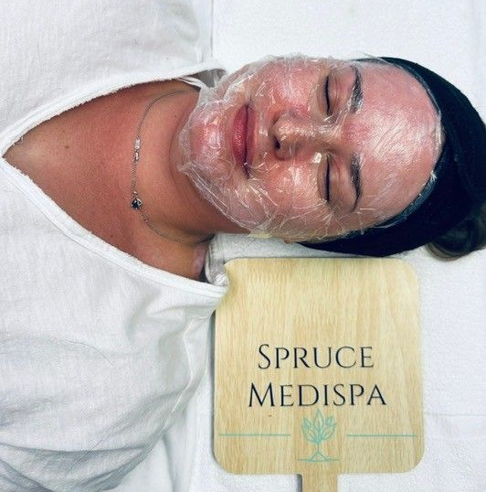 A person lying down with a plastic film mask on their face, holding a wooden paddle with