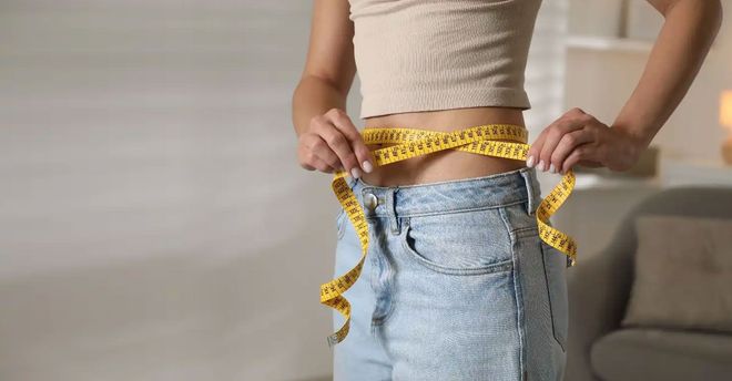 Person measuring their waist with a yellow tape measure, wearing jeans and a crop top indoors.