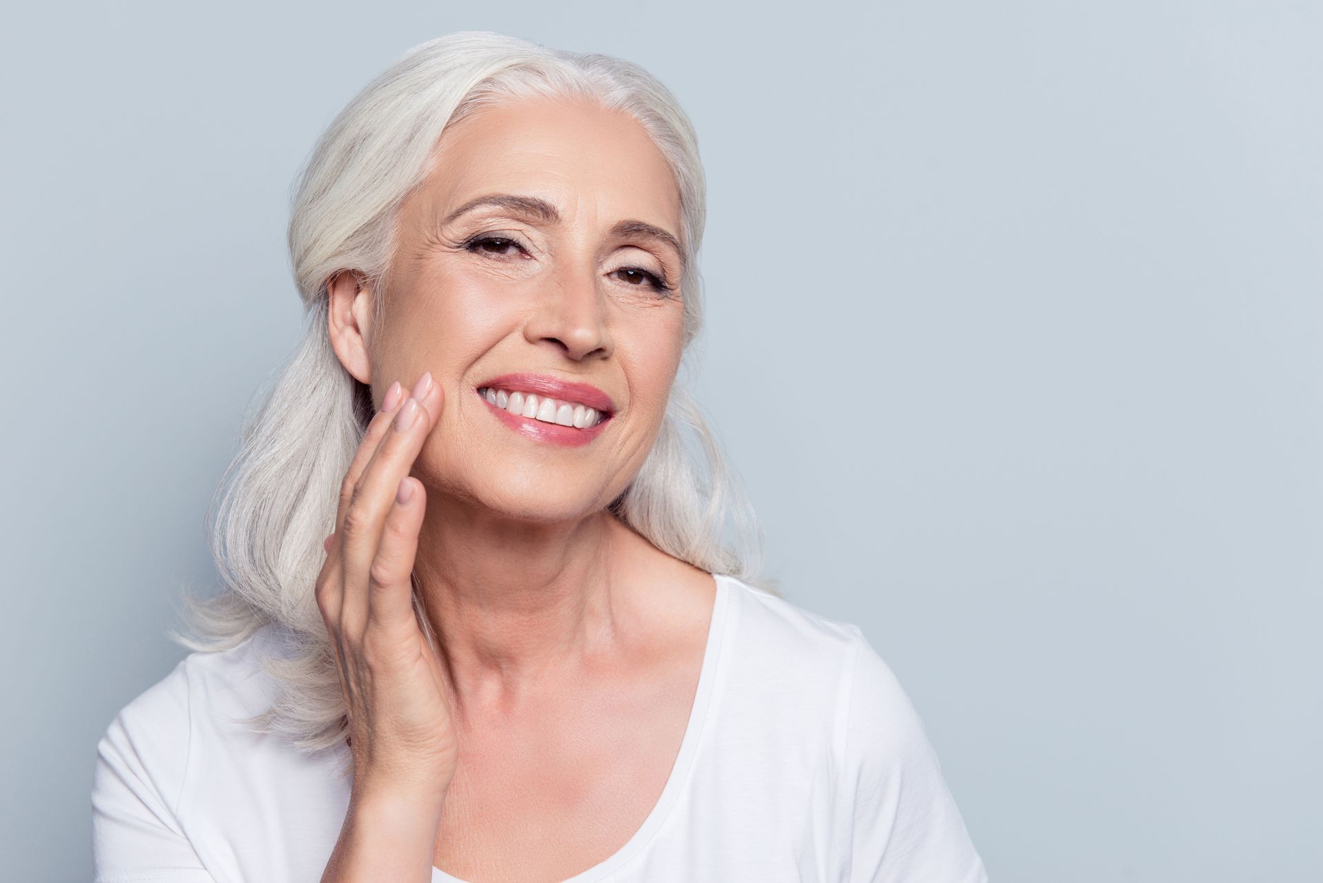 Smiling woman with white hair touching her cheek against a light gray background