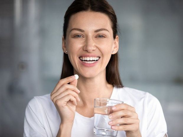Smiling woman holding a pill and a glass of water in a bright indoor setting