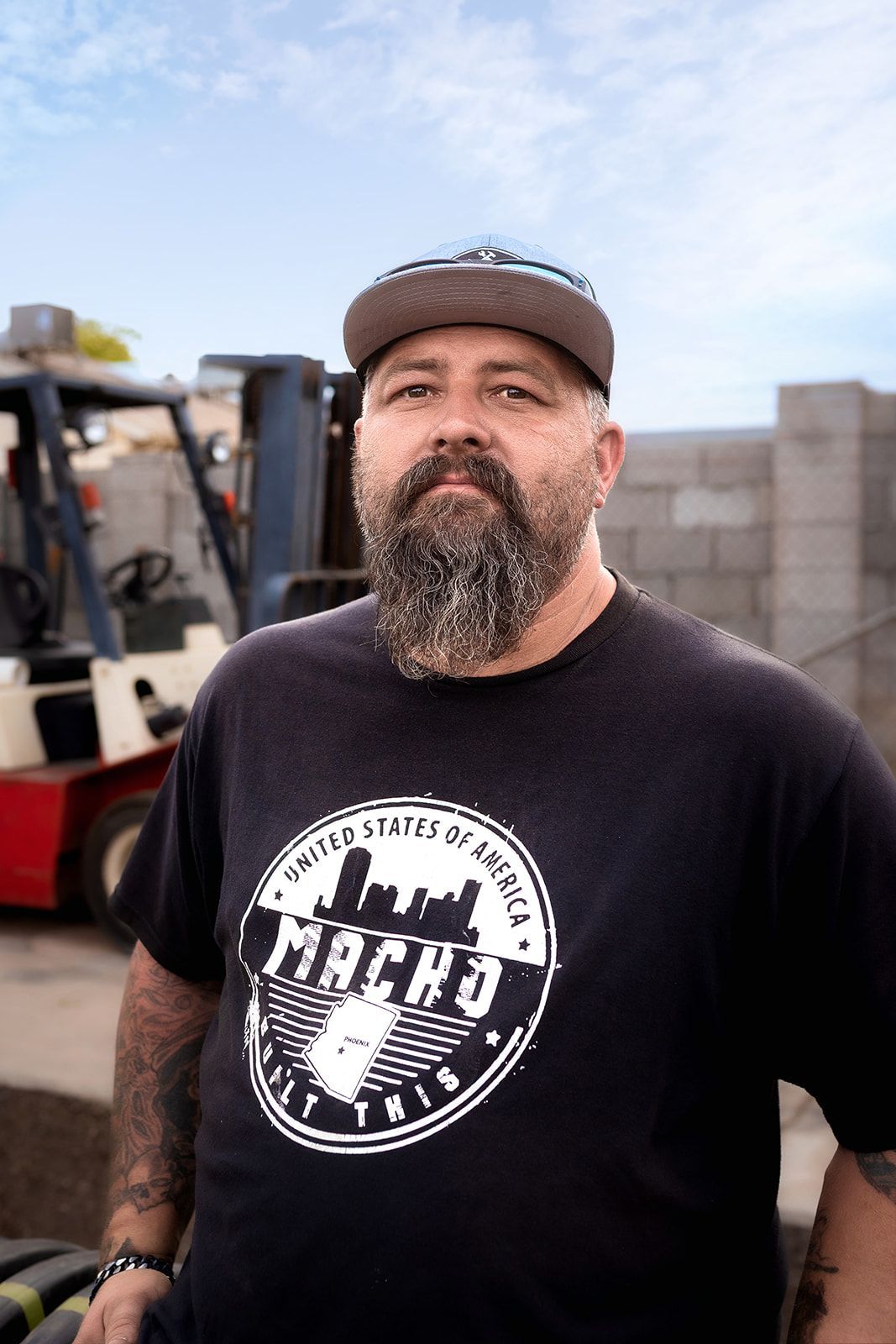 A man with a beard and a hat is standing in front of a forklift.