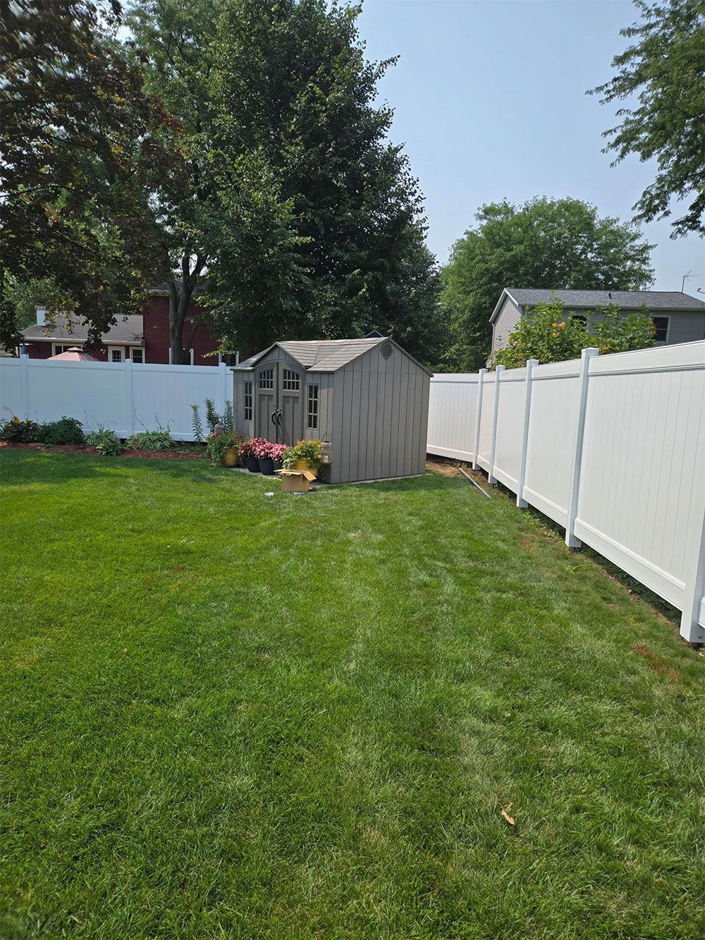 Backyard with white fence, shed, green grass, and trees. Sunny day.
