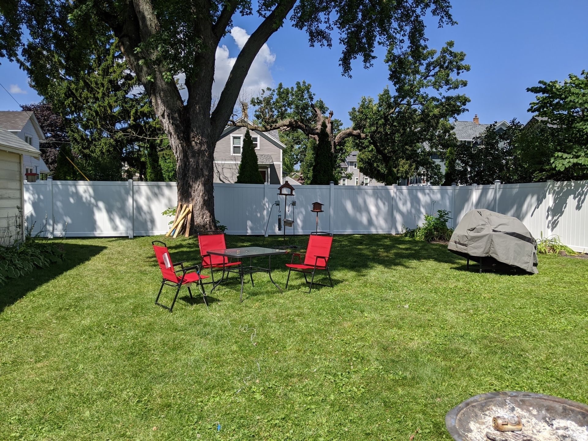 Backyard with white fence, red chairs around a table, large tree, and a boulder on a sunny day.