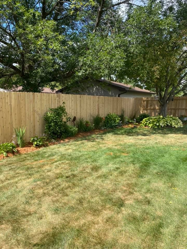 Wooden fence bordering a backyard with grass, flower beds, and trees on a sunny day.