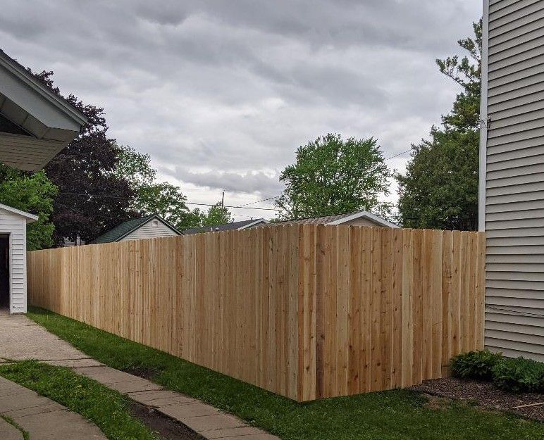 Wooden fence surrounds a yard with grass, a driveway, and houses under a cloudy sky.