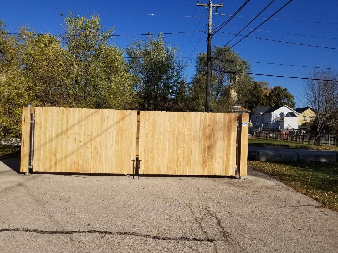 Wooden gate closed across a paved road, trees and houses in the background under a blue sky.