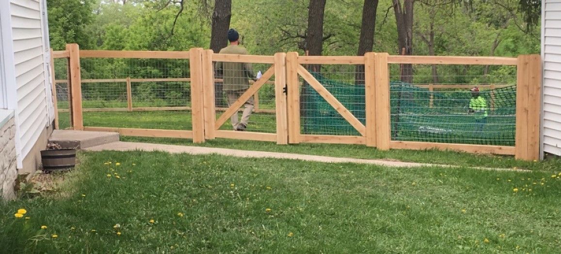 Wooden fence with two gates, enclosing a yard with green grass. A person stands behind the open gate.