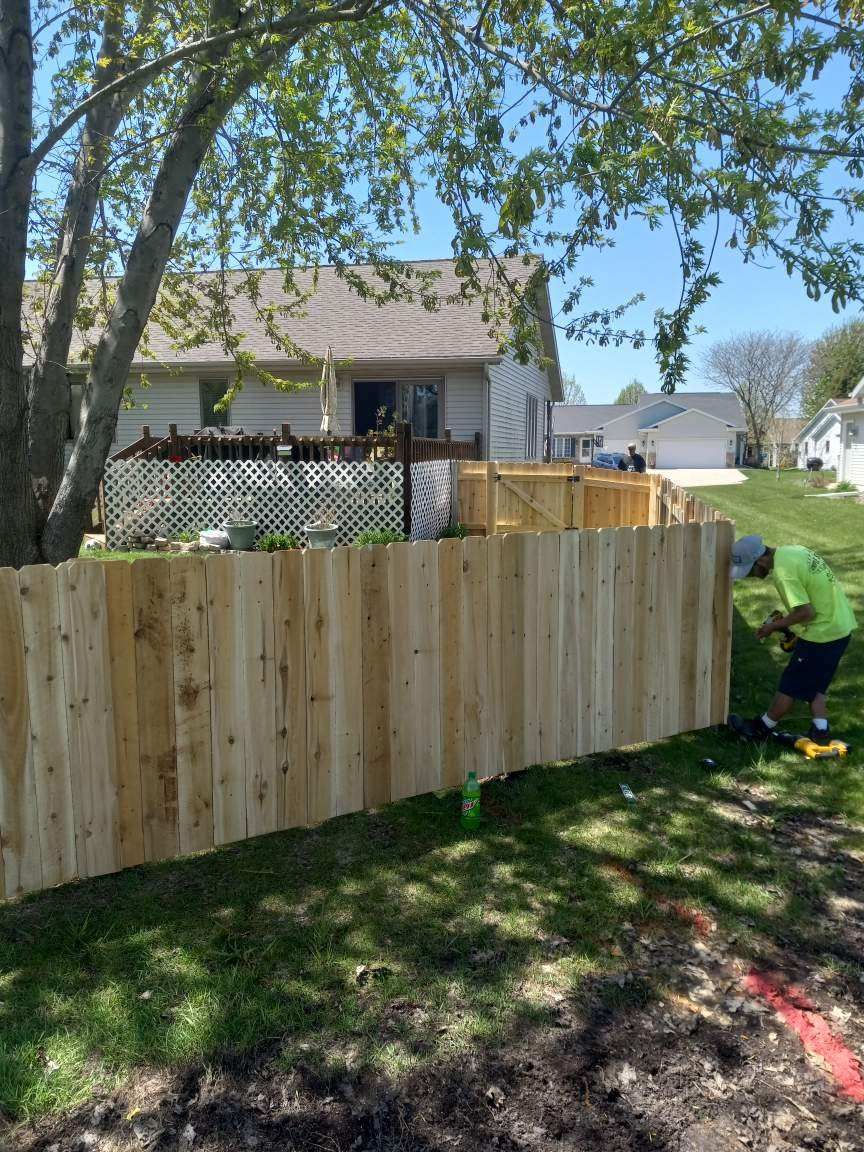 Person installing a new wooden fence in a yard with a house in the background on a sunny day.