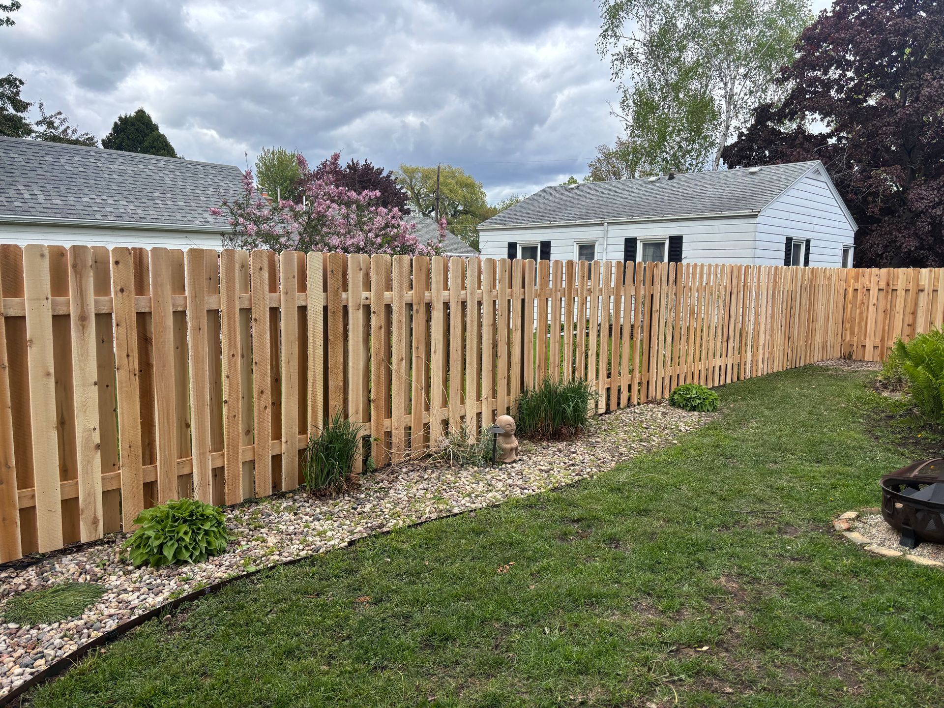 Wooden picket fence curves along a backyard, with a stone border and green grass. Houses in the background.