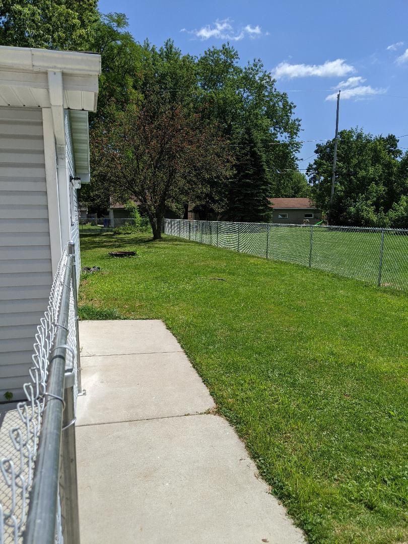 Side view of a house with a concrete walkway, green grass, a chain-link fence, and a tree under a blue sky.
