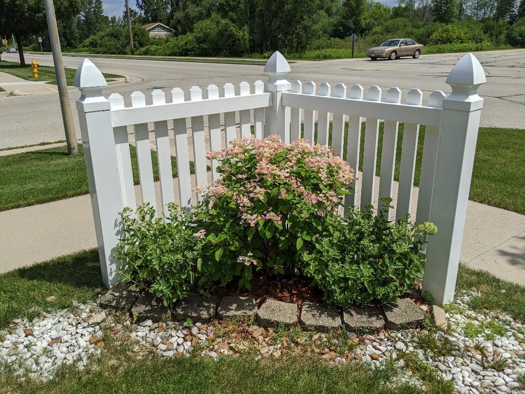 White picket fence encloses a small garden with green and pink plants, set in gravel and bordered by stone.