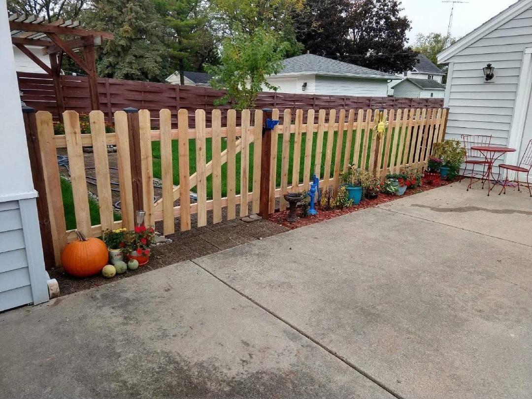 Wooden picket fence with a gate, lined with potted plants and pumpkins next to a house with a concrete patio.