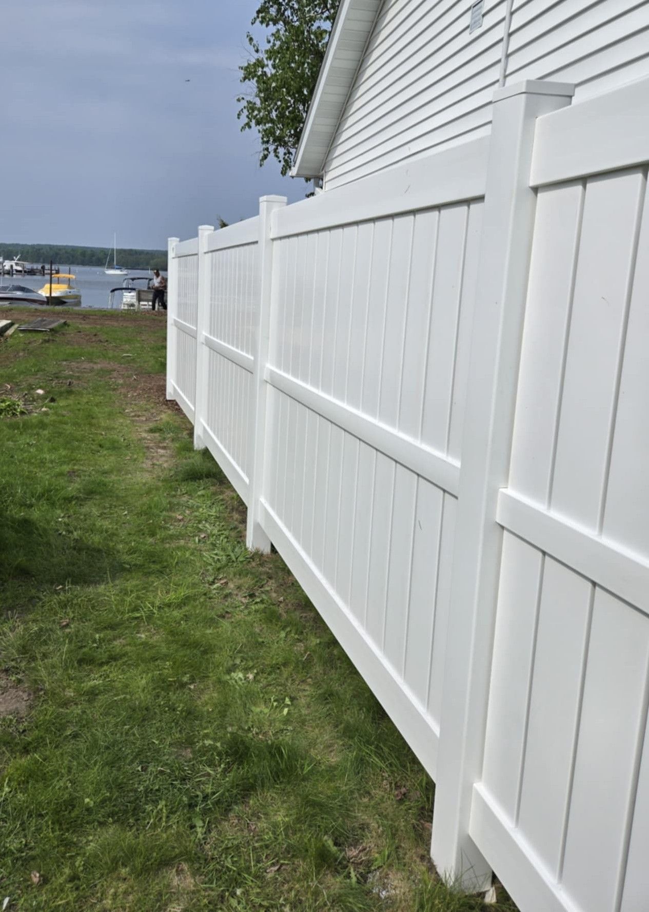 White vinyl fence alongside a building and green grass, with a body of water in the background.