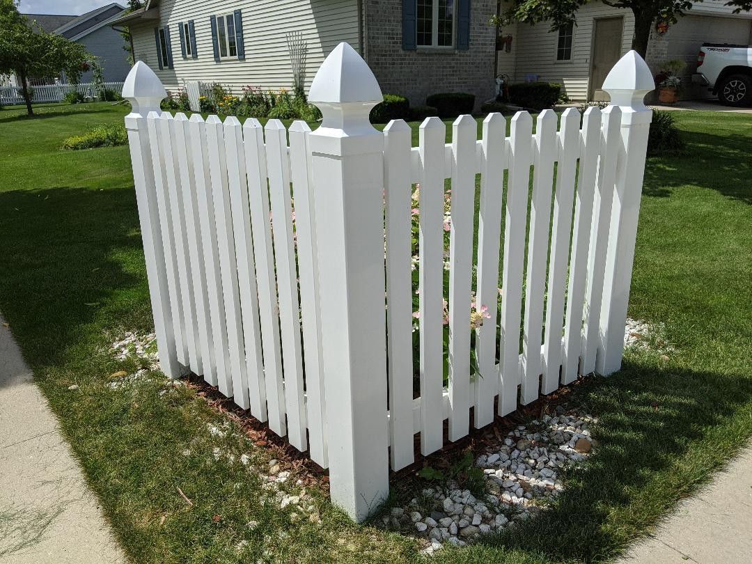 White picket fence surrounding a small tree in a grassy yard, near a sidewalk and house.