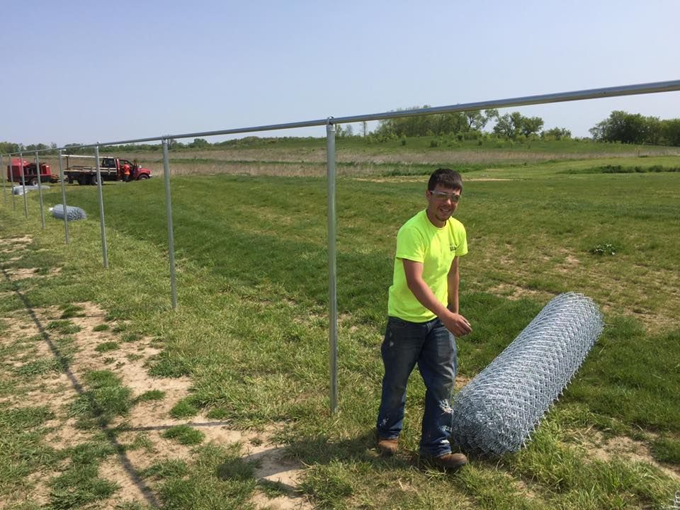Person rolling out wire fencing next to a partially built metal fence in a grassy field.