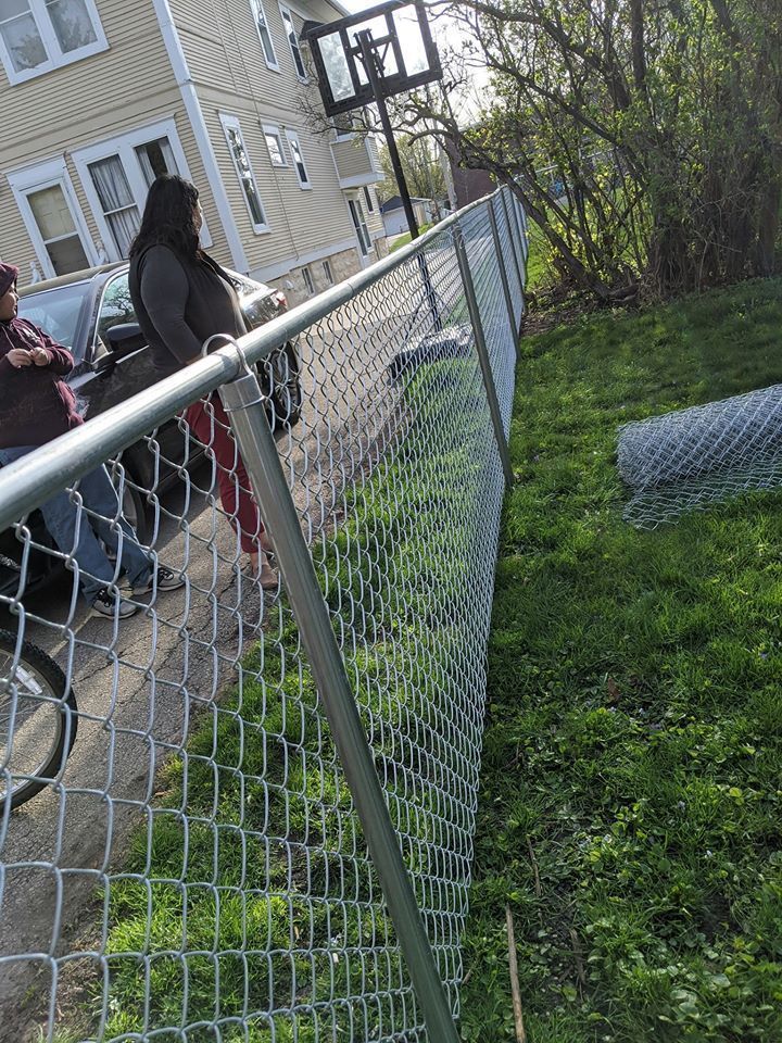 Chain-link fence in front yard with two people near a car; a basketball hoop is visible in the background.
