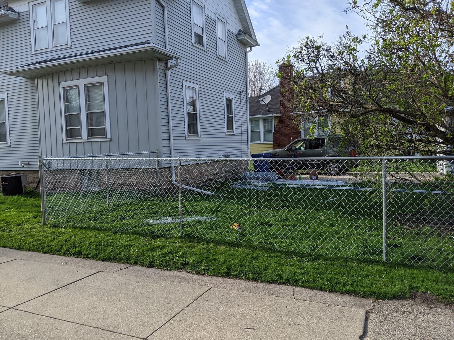 Chain-link fence in front of a gray house with a lawn; a car and another house are visible in the background.