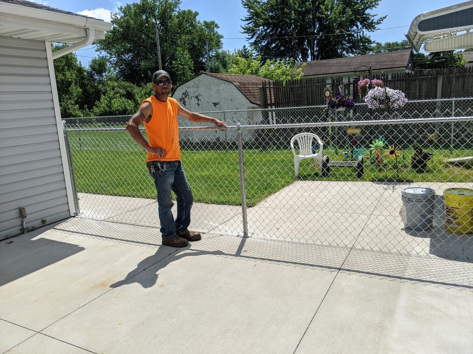 Man in orange shirt leaning on chain-link fence in a backyard with a concrete patio.