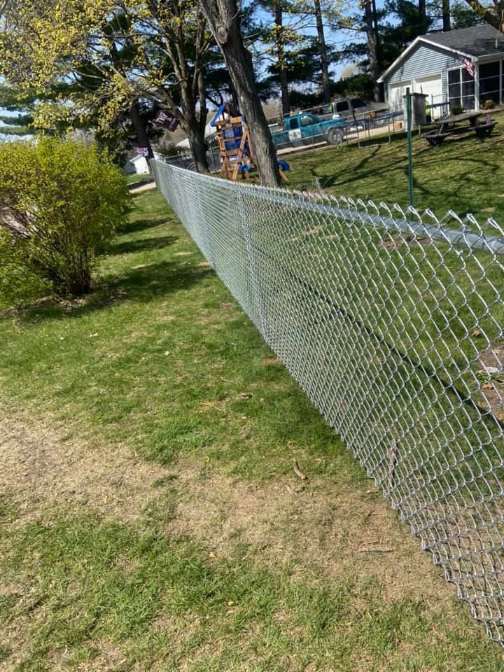 Chain-link fence on a grassy lawn, trees and a house in the background on a sunny day.