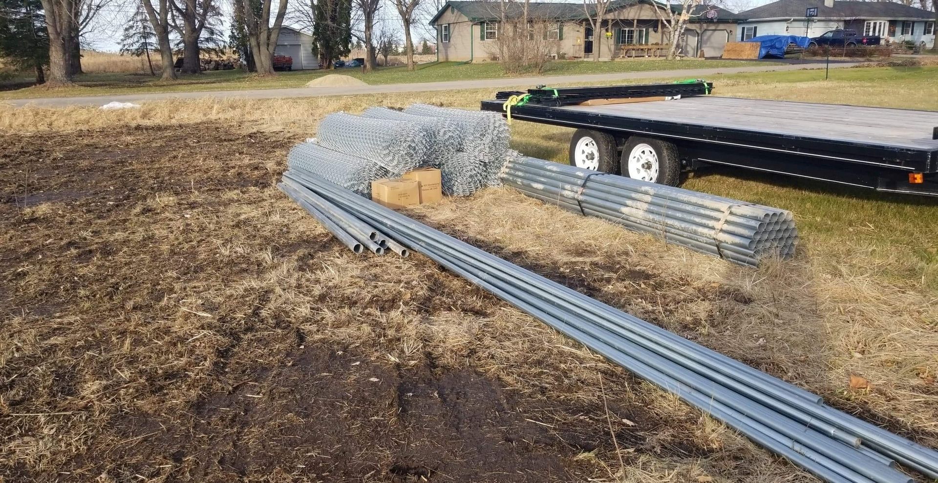 Metal fence components are on the ground near a trailer in a field. Brown and green landscape.