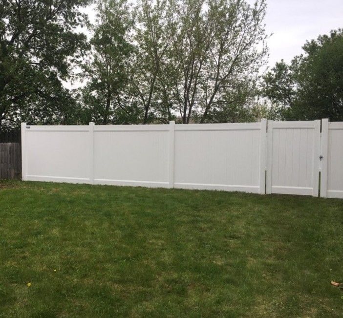 White vinyl privacy fence in a grassy yard, with a gate and trees in the background.