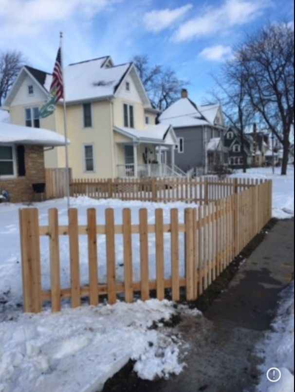 Wooden picket fence in snowy yard, houses in background, American flag on pole.