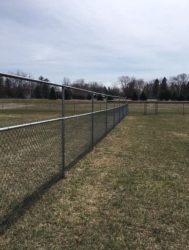 Chain-link fence in a grassy field on a cloudy day, with trees in the background.