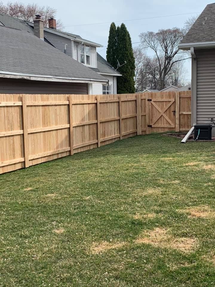 Wooden fence encloses a green lawn with a gate, adjacent to houses with gray roofs.