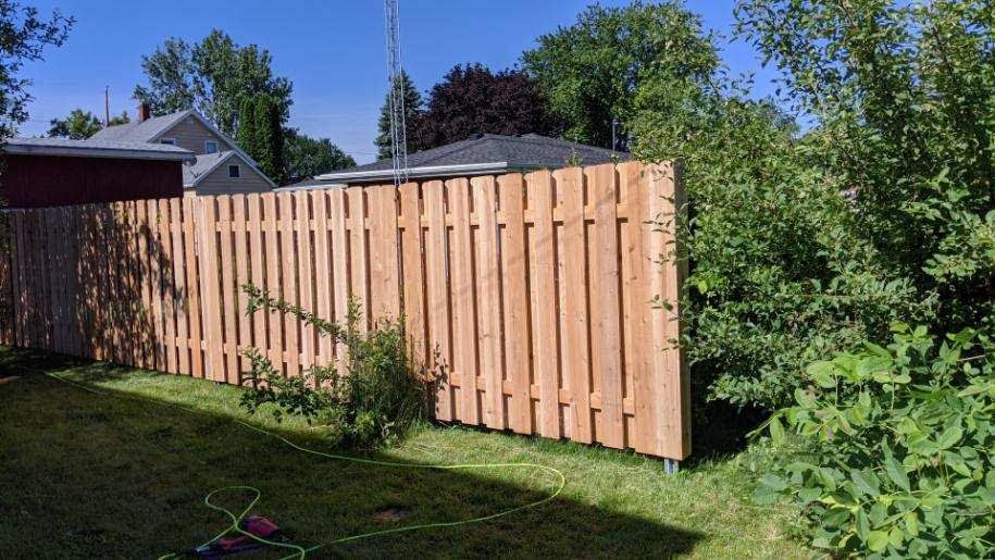 Wooden fence in a grassy yard, partially obscuring a house and bushes. Bright sunlight.