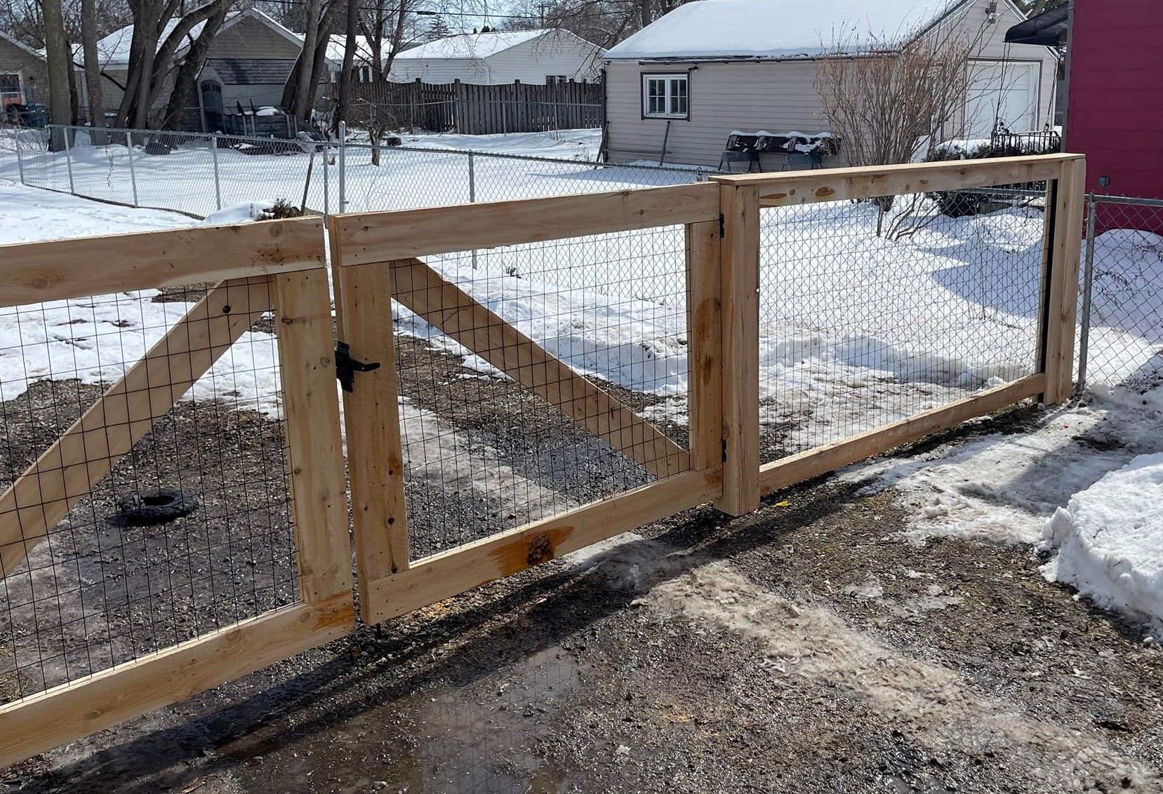 Wooden gate with wire mesh, in a snowy gravel yard, attached to chain-link fence.