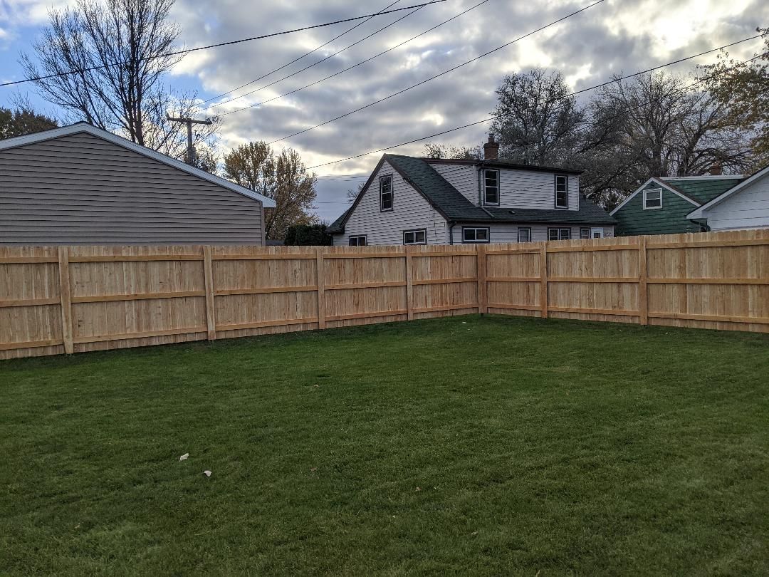 Wooden fence encloses a green lawn in a suburban backyard, houses visible in the background under a cloudy sky.