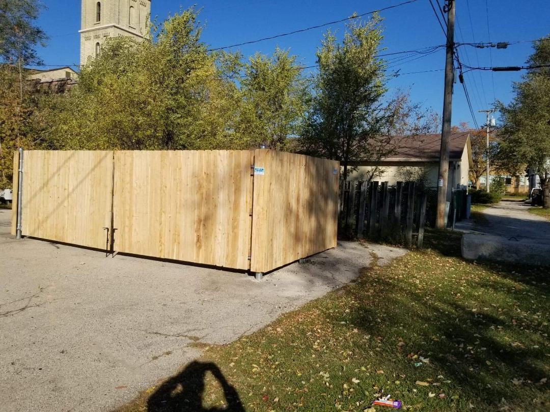 A wooden fence blocks a driveway, with trees and a building in the background. Sunny day.