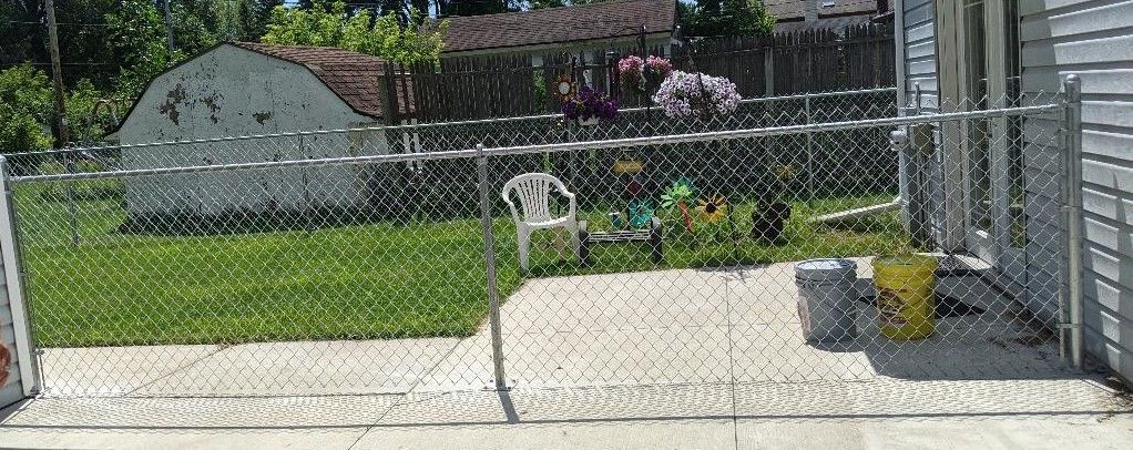 Chain-link fence encloses a yard with grass, a patio, and a white chair. Trees and a house are in the background.
