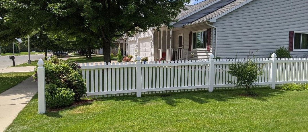 White picket fence in front of a house. Lush green grass and trees surround the house. Sunny day.