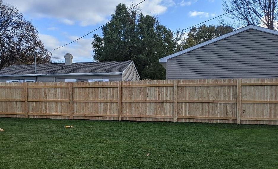 Wooden fence in a backyard with a grassy lawn, with houses and blue sky in the background.