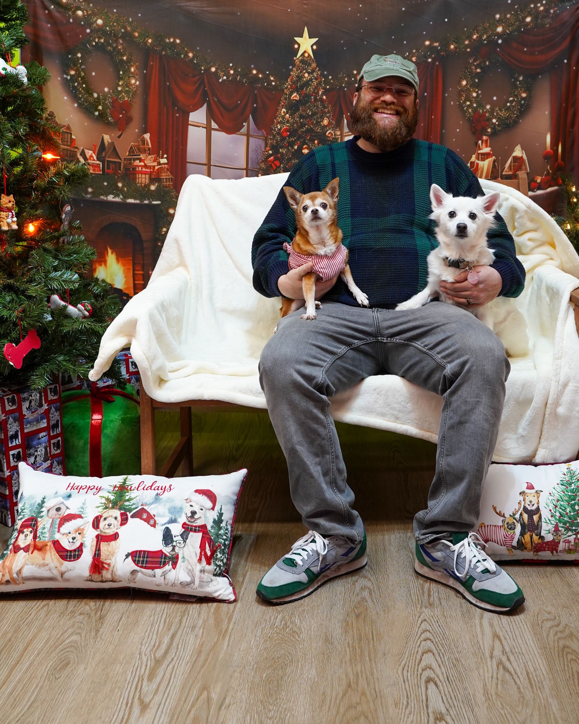 Man seated with two dogs in front of a holiday backdrop. He wears a sweater and hat, holding the dogs. Festive pillows on floor.