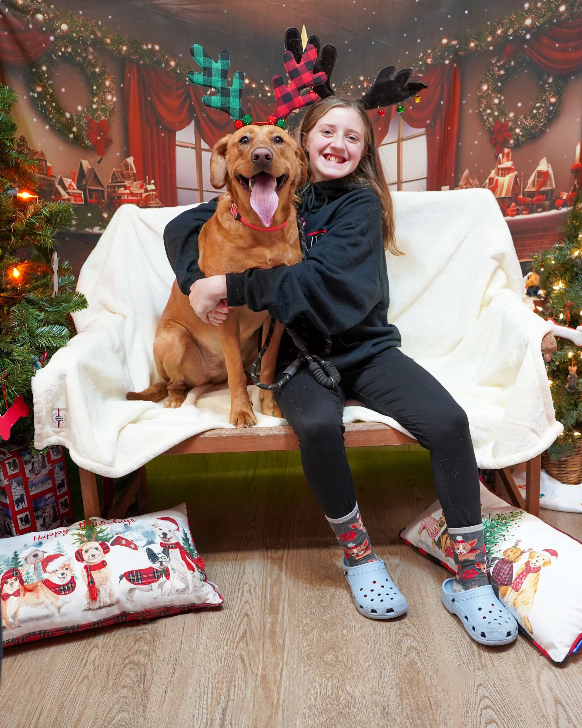 Girl with brown dog wearing reindeer antlers, smiling, in front of a Christmas backdrop.