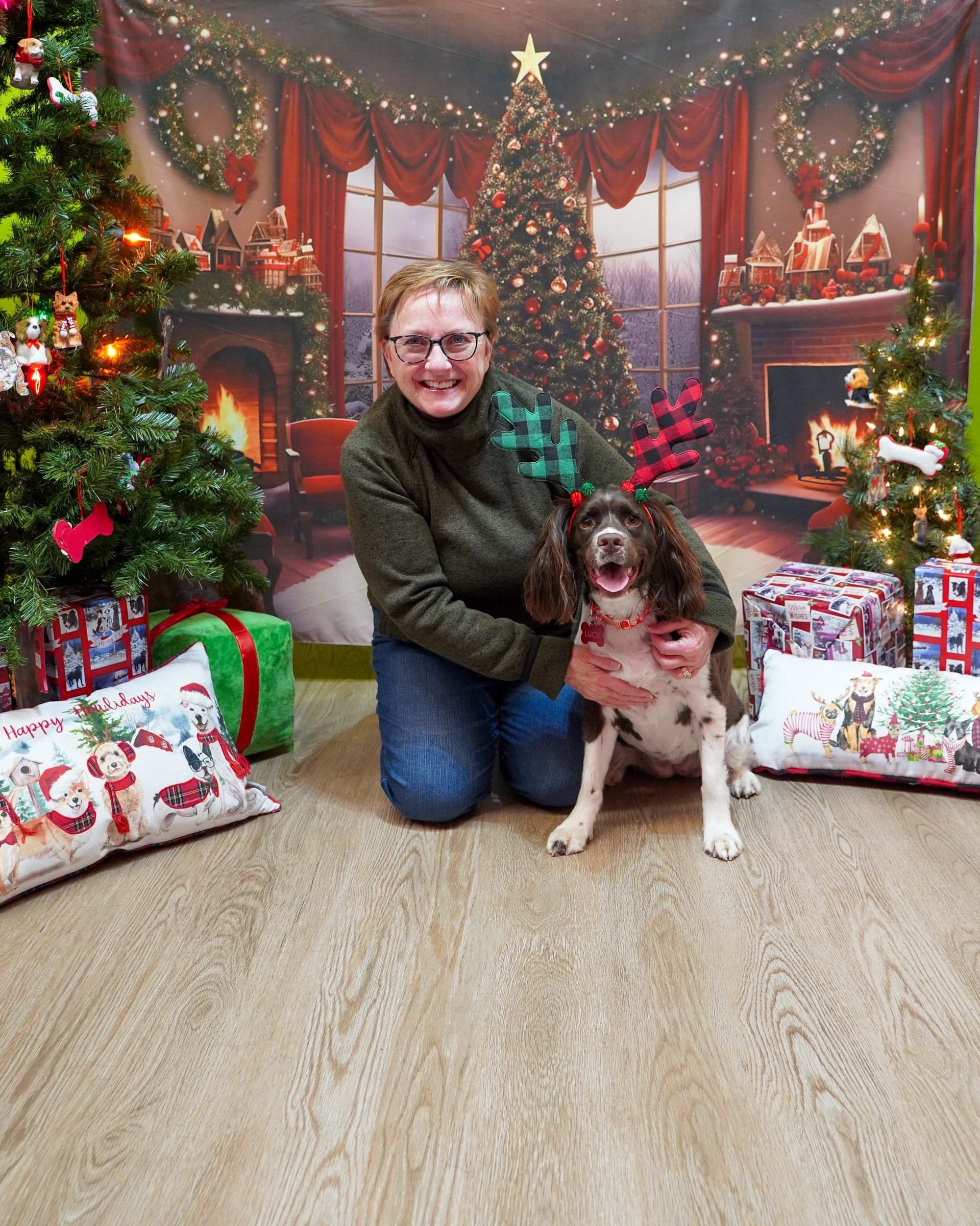 Woman kneeling with a brown and white dog wearing reindeer antlers, posing in front of a Christmas backdrop.