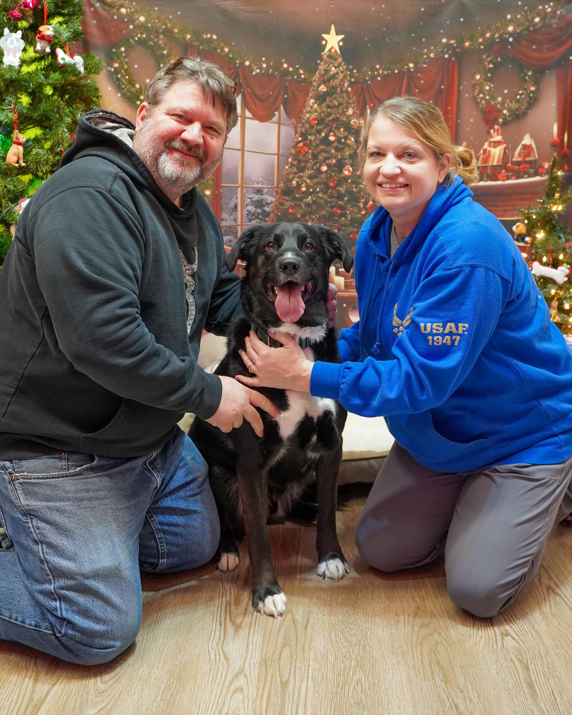 Couple kneeling with a black and white dog in front of a Christmas backdrop. Dog is smiling with its tongue out.