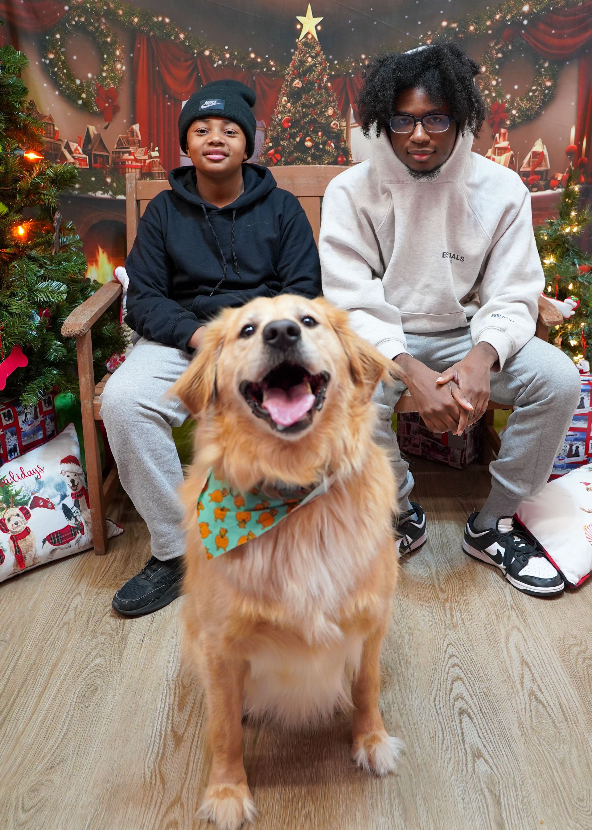 Two people and a golden retriever pose for a Christmas photo. The dog smiles with a bandana. They sit in front of a Christmas backdrop.